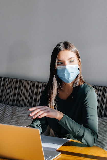 Een jong meisje zit in een café met een masker op en werkt op een computer.