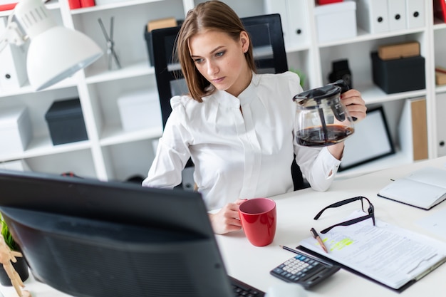 Een jong meisje zit op een computer Bureau in het kantoor en giet zijn koffie.