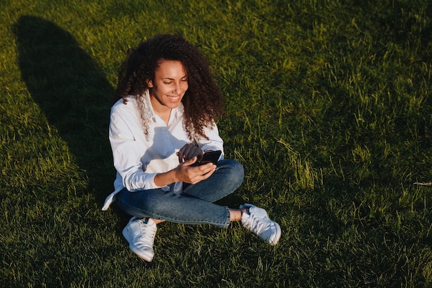 Een jong meisje zit op het groene gras in het park en gebruikt sociale netwerken met een mobiele telefoon