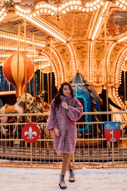 Een jong mooi meisje in een bontjas staat op een winteravond bij de carrousel