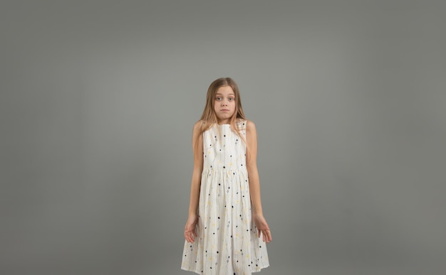 Een jong mooi meisje in een lichte jurk haalt haar schouders op tegen een grijze geïsoleerde achtergrond.