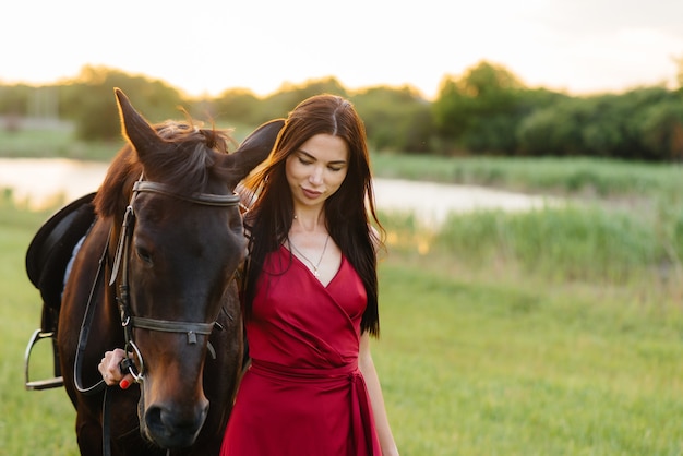 Een jong mooi meisje in een rode jurk vormt bij zonsondergang op een boerderij met een volbloedhengst. Liefde en zorg voor dieren.