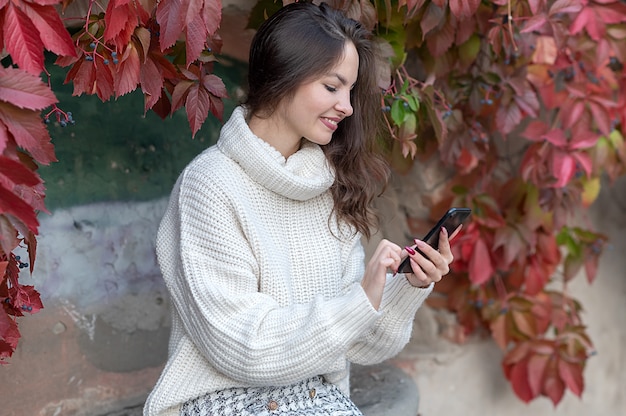 Een jong mooi meisje in een witte trui kijkt naar de telefoon. In de herfst op straat.
