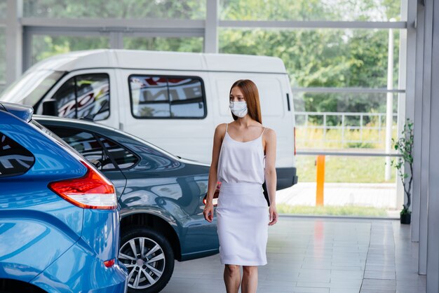 Een jong mooi meisje inspecteert een nieuwe auto bij een autodealer in een masker tijdens de pandemie. De verkoop en aankoop van auto's, in de periode van pandemie.