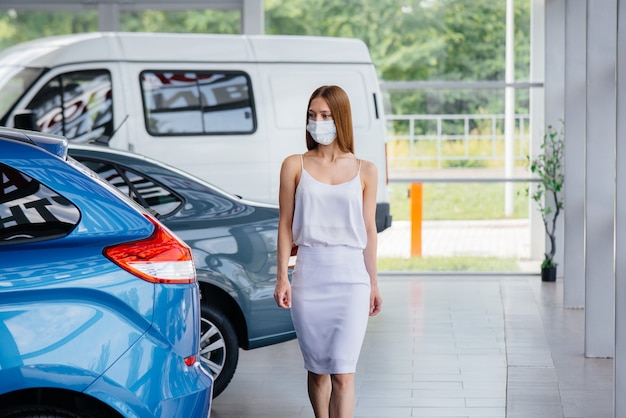 Een jong mooi meisje inspecteert tijdens de pandemie een nieuwe auto bij een autodealer in een masker. De aan- en verkoop van auto's, in de periode van pandemie.