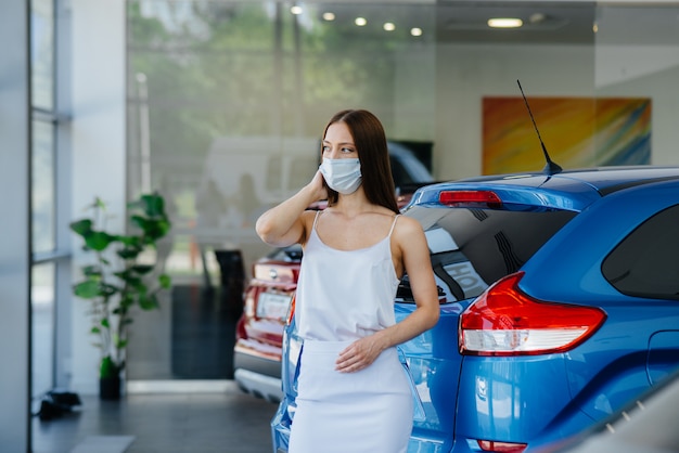 Een jong mooi meisje inspecteert tijdens de pandemie een nieuwe auto bij een autodealer in een masker. De aan- en verkoop van auto's, in de periode van pandemie.