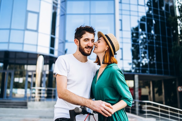 Een jong romantisch koppel met elektrische scooters op een date, wandelen in de stad. Jonge vrouw in hoed en man geniet van een wandeling