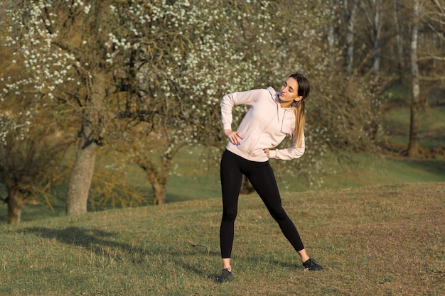 Een jong slank atletisch meisje in sportkleding voert een reeks oefeningen uit. Fitness en gezonde levensstijl tegen de achtergrond van groene heuvels van de lenteweide.