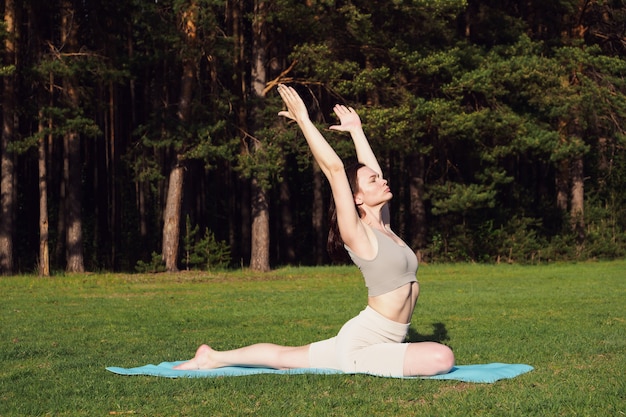 Een jong slank meisje doet yoga, doet asana's op een mat, in de natuur. buitensporten, gezond levensstijlconcept. een vrouw brengt een fitnesssessie door in het park, in het bos, in de zon.