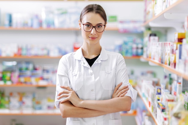 Een jong slank meisje met lang donker haar en een bril, gekleed in een witte jas, staat zelfverzekerd bij de planken in een up-to-date apotheek. .