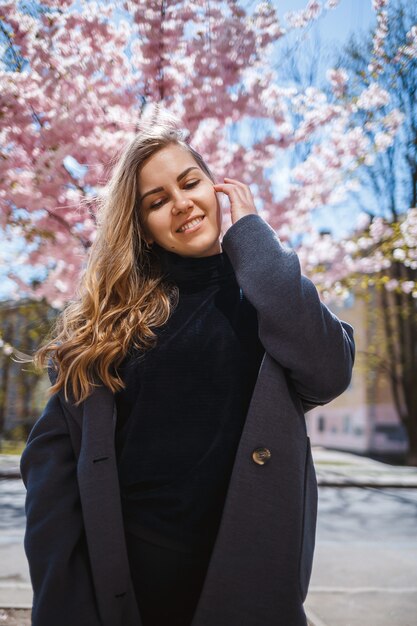 Een jong slank vrouwelijk model met lang golvend haar en gekleed in een grijze jas, sneakers, staat op straat bij een bloeiende struik met prachtige roze bloemen op de achtergrond en poseert.