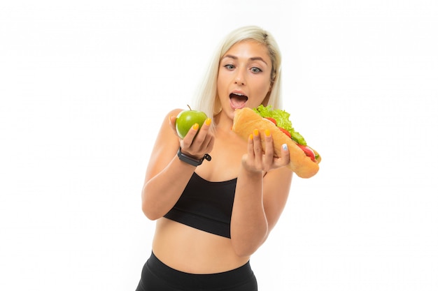 Een jong sportmeisje met blond haar in een zwarte sporttop en zwarte legging heeft een groene appel en sandwich.