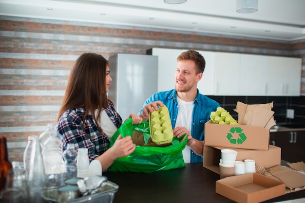 Een jong stel sorteert afval in de keuken. De jonge man en de vrouw sorteren recycleerbare voorwerpen in de keuken. Er is karton, papier, ijzer, plastic en glas en andere materialen die gerecycled kunnen worden.