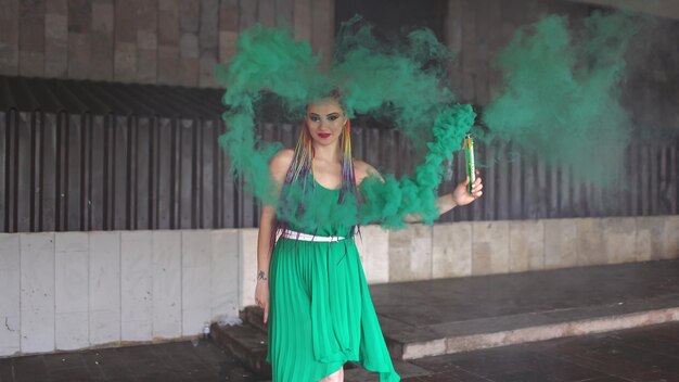 Een jong vrolijk meisje in een felgroene lentejurk met make-up met pailletten en regenboog afrikaanse vlechten. ze staat bij een gebouw en blaast groene rook op straat.