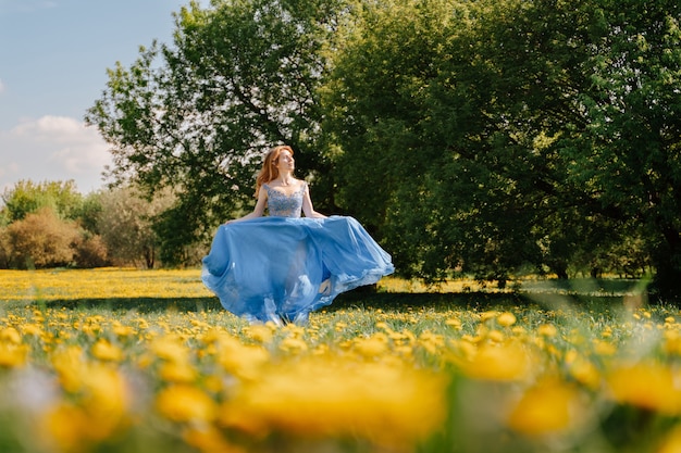 Een jong vrolijk meisje rent in een blauwe lange jurk over een veld met gele weidebloemen, een zomergazon met paardebloemen