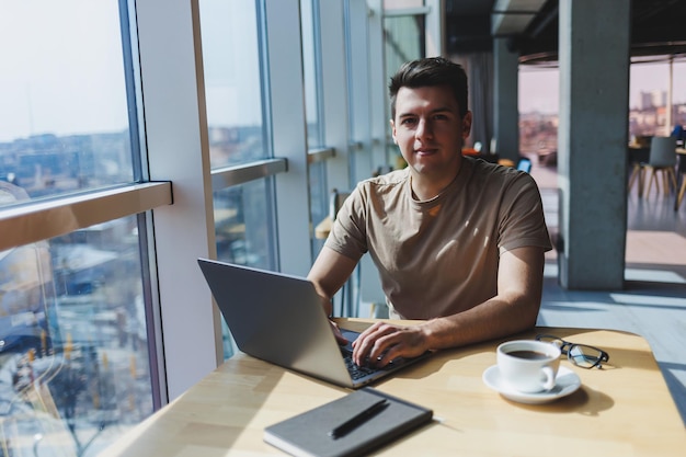 Een jonge aantrekkelijke mannelijke freelancer zit in het interieur van een coffeeshop en kijkt uit het raam Knappe blogger schrijft ideeën in laptop zittend aan houten tafel in café met laptop