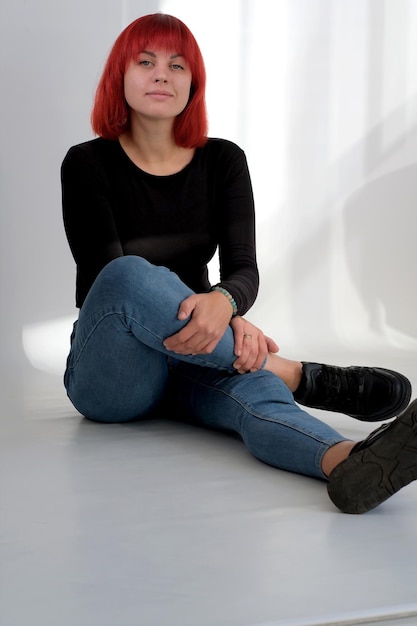 Een jonge aantrekkelijke vrouw met een kort oranje kapsel in een zwart T-shirt en spijkerbroek poserend in de studio op een witte achtergrond