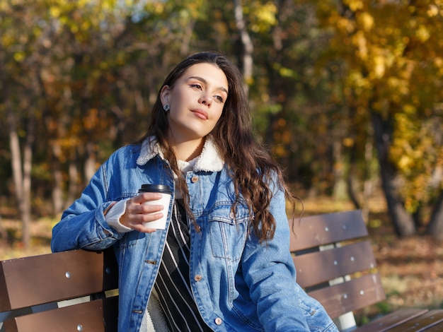 Een jonge aantrekkelijke vrouw zit op een bankje in een herfstpark met een kopje koffie