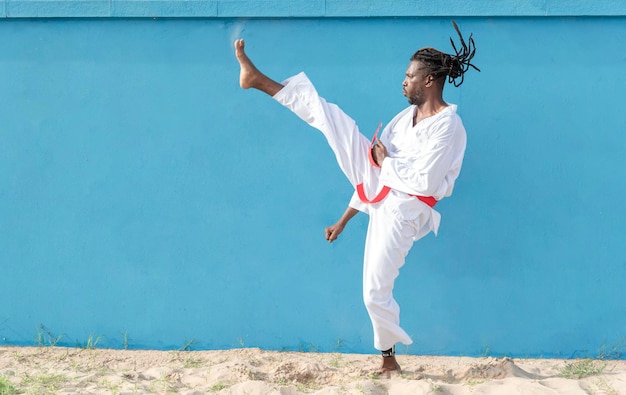 Een jonge afro-amerikaanse man die taekwondo buiten traint en schopt op een blauwe achtergrond vechtsportconcept