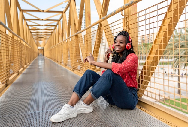 Een jonge Afro-Amerikaanse vrouw in vlechten die op een gele brug zit en naar muziek luistert met haar rode koptelefoon en mobiele telefoon