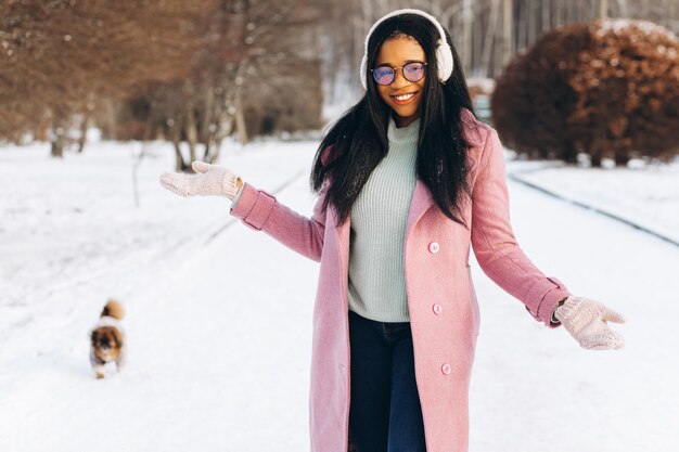 Een jonge Afro-Amerikaanse vrouw met een bril en warme wanten laat haar hond uit in een winterpark