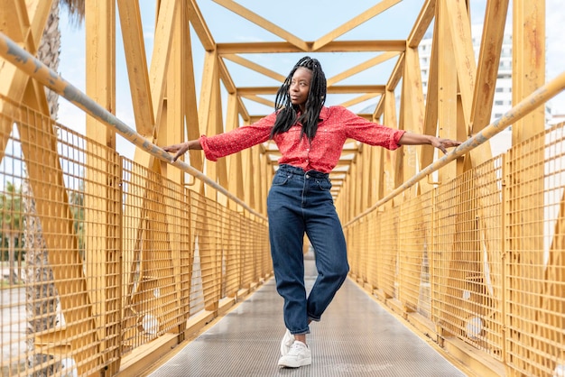 Een jonge Afro-Amerikaanse vrouw met vlechten die op een gele brug staat met uitgestrekte armen op de reling