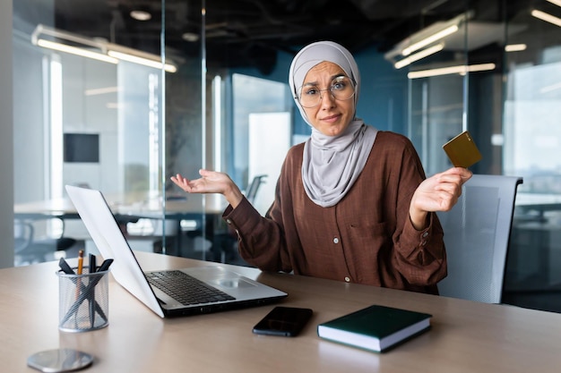 Een jonge Arabische vrouw die op kantoor aan een tafel zit met een laptop en een creditcard in haar hand houdt