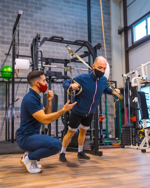 Een jonge atleet met een instructeur in de sportschool doet oefeningen met strips in de pandemie van het coronavirus