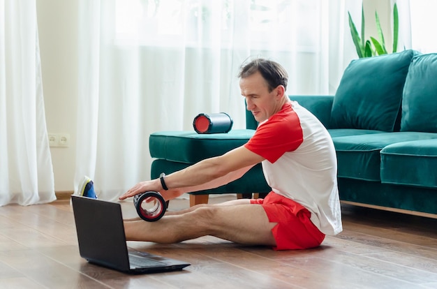 Een jonge atletische man in rode korte broek doet oefeningen op een schuimroller met behulp van een notebooklaptop. Werkt uit de spieren met een massageroller. Online sporten of video-tutorial volgen