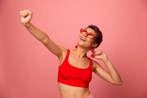 Een jonge atletische vrouw met een kort kapsel en paars haar in een rode top in zonnebril met een atletische figuur glimlacht met haar armen uitgestrekt in verschillende richtingen dansend op een roze achtergrond