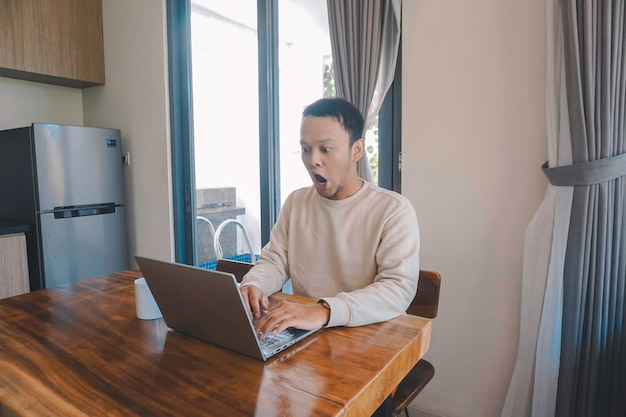 Een jonge Aziatische man brengt zijn tijd thuis door terwijl hij geschokt in de eetkamer zit terwijl hij naar zijn laptop kijkt