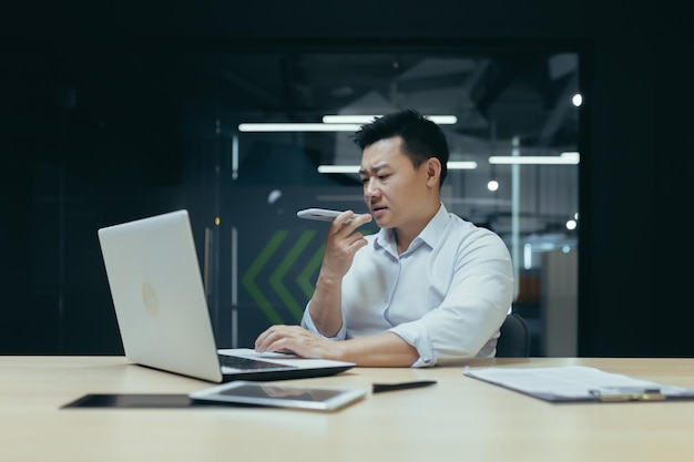 Een jonge aziatische man zit op kantoor aan tafel en werkt aan een laptop die aan de telefoon praat