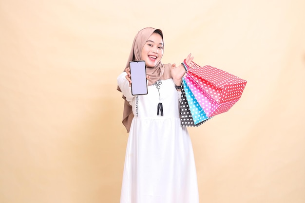 een jonge Aziatische moslimvrouw in een gelukkige hijab toont een mobiele telefoon scherm en draagt een paperbag shop
