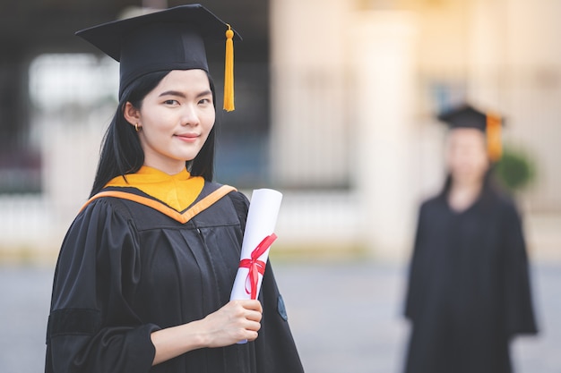 Een jonge Aziatische vrouw afgestudeerd aan de universiteit in afstudeerjurk en baret heeft een diploma staat voor het universiteitsgebouw na deelname aan het begin van de universiteit