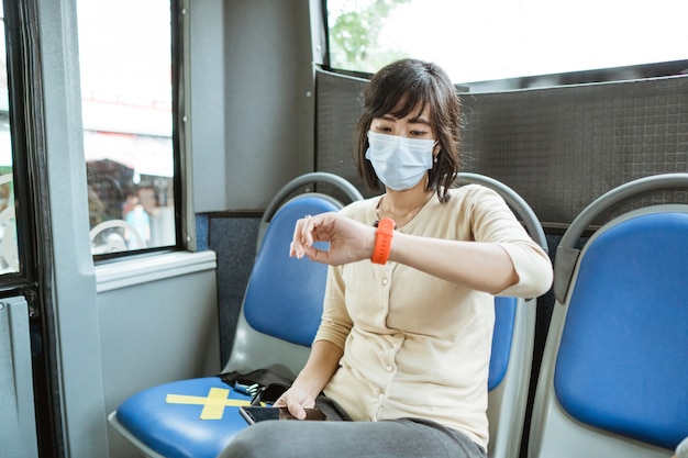 Een jonge Aziatische vrouw die een masker draagt, zit op een bank tijdens het reizen naar haar horloge in de bus te kijken