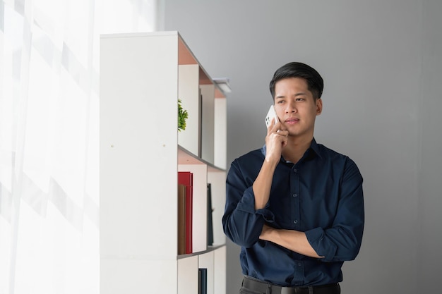 Een jonge Aziatische zakenman staat op het kantoor van een raam en praat aan de telefoon