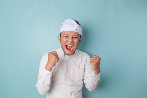 Een jonge Balinese man met een gelukkige succesvolle uitdrukking die udeng of traditionele hoofdband en wit overhemd draagt, geïsoleerd door blauwe achtergrond