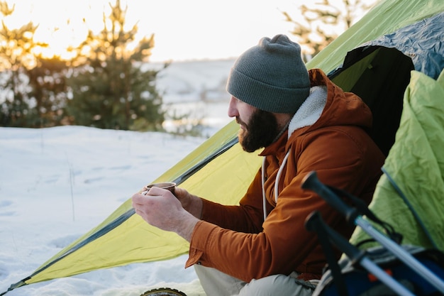 Een jonge, bebaarde man rust in de winterbergen bij een tent Een mantraveler met een baard in een muts en een warme jas warmt op door hete thee of koffie te drinken na een wandeling