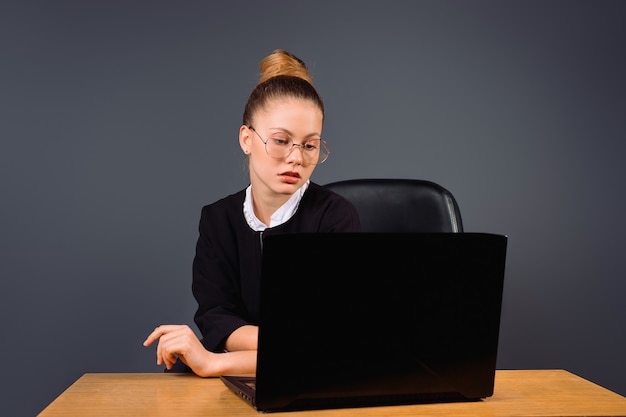 Een jonge bedrijfsvrouw met bril zit bij een werkende computerfoto op een grijze geïsoleerde achtergrond