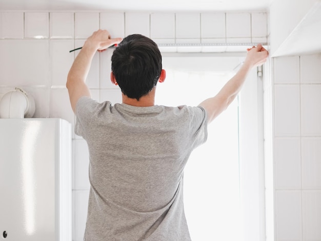 Een jonge blanke man meet de breedte van een klein raam in de badkamer met een tape