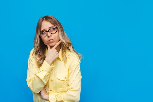 Een jonge blanke mooie blonde doordachte student vrouw in een geel shirt denkt weg te kijken naar lege kopieerruimte voor advertenties promo aanbieding citaat tekst slogan geïsoleerd op een felle kleur blauwe muur