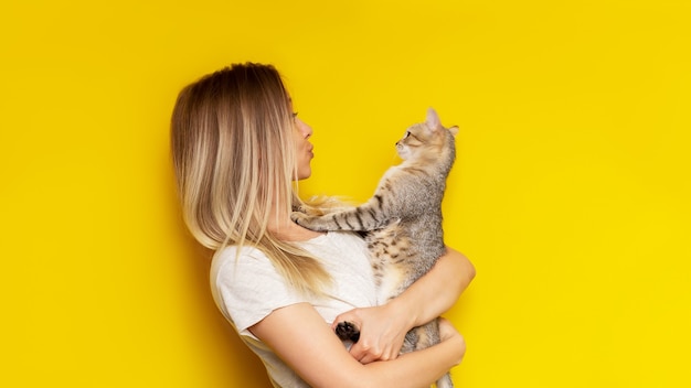 Een jonge blanke mooie schattige blonde vrouw houdt een gestreepte kat in haar handen en bewondert het geïsoleerd op een felle gele muur het meisje babying met een kitten fiendship van huisdier en eigenaar