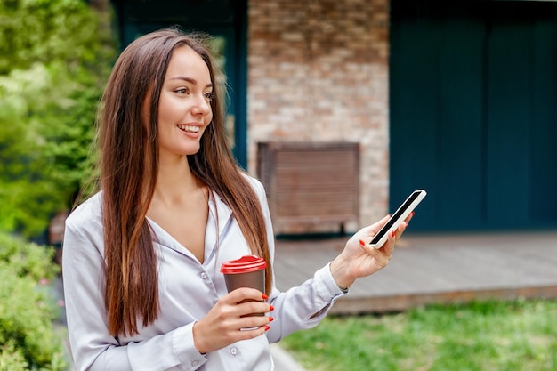 Een jonge blanke vrouw gebruikt haar smartphone, houdt een papieren kopje koffie vast en lacht op straat