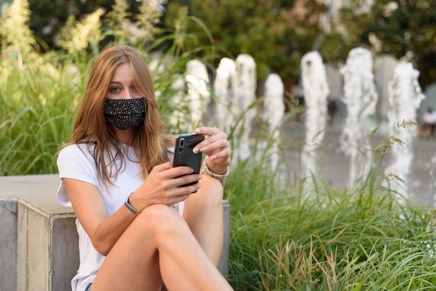 Foto een jonge blanke vrouw met een masker kijkt door haar telefoon in een straat