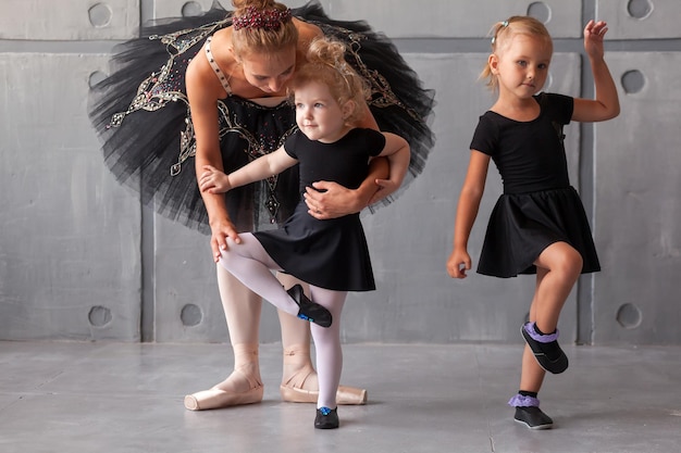 Een jonge blonde ballerina in een zwarte feestelijke jurk, witte panty en spitzen leert twee kleine onoplettende meisjes in zwarte jurken om een Russisch ballet te dansen in een donkere dansstudio
