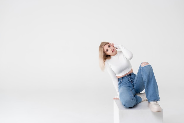 Een jonge blonde in een witte top en jeans zit op een witte kubus en poseert op een afgelegen witte achtergrond in de studio. Mensen levensstijl concept. Kopieer ruimte voor kopiëren