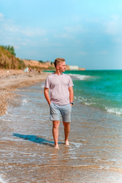 Een jonge blonde man loopt op het strand in de buurt van de azuurblauwe oceaan of zee Het concept van een gelukkige zomer