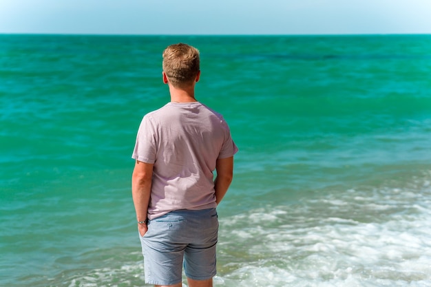 Een jonge blonde man loopt op het strand in de buurt van de azuurblauwe oceaan of zee Het concept van een gelukkige zomer