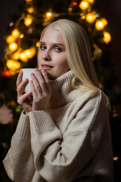 Een jonge blonde vrouw drinkt geurige cacao thuis in de buurt van een kerstboom.