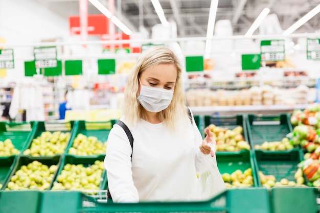 Een jonge blonde vrouw met een medisch masker koopt fruit in een supermarkt